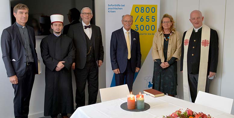 Ein Foto von sechs Personen, die sich in einem Raum des Krisendienstes Psychiatrie Oberbayern befinden. Vor Ihnen ist ein Tisch mit einer weißen Tischdecke, vier Kerzen und Blumen zusehen.
