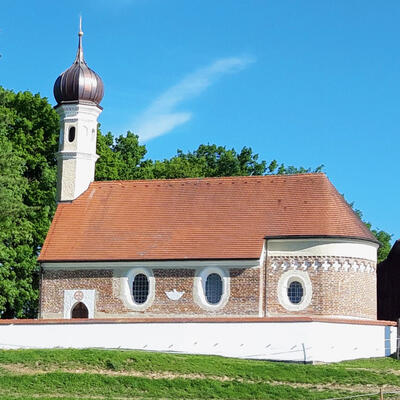 Seitenansicht einer kleinen Kirche aus Zielgelsteienen mit einem weißen Zwiebelturm. Umgeben ist die Kircche von eienr weißen Mauer.